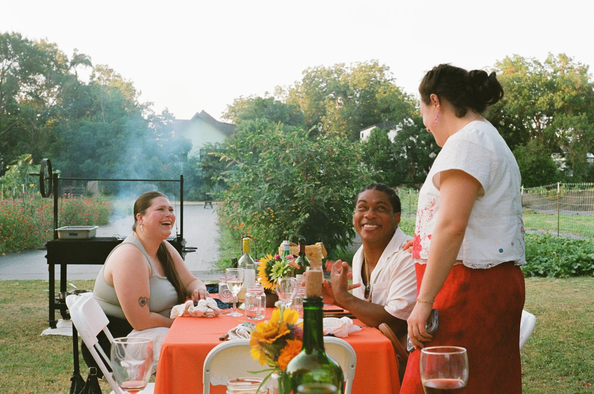Friends laughing around an outdoor table at golden hour, a live-fire grill smoking in the background